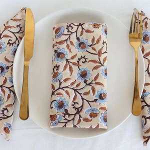 May include: Three beige cotton napkins with a blue and brown floral pattern are folded and placed on a white plate with gold silverware.