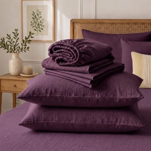Peut inclure: Un ensemble de linge de lit violet profond comprenant un drap-housse, un drap plat et deux taies d'oreiller rectangulaires. Le tissu présente une texture tissée visible. Les articles sont empilés proprement sur un lit, affichant une couleur prune unie.