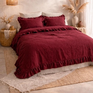 May include: A bed dressed with a deep red duvet cover and pillowcases, all with a ruffled trim. The bed sits on a neutral rug. A woven pendant light hangs above the bed, creating a warm atmosphere.