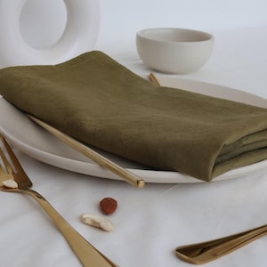 May include: A folded olive green linen napkin on a white plate with gold cutlery. The napkin is folded in a simple, elegant way, and the cutlery is arranged neatly beside it.