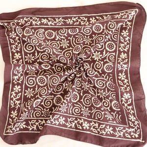 May include: A dark brown square scarf with a white floral and swirl pattern. The scarf is folded and has a decorative border. The scarf is made of a soft material and is suitable for various occasions.