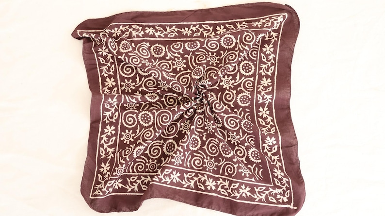 May include: A dark brown square scarf with a white floral and swirl pattern. The scarf is folded and has a decorative border. The scarf is made of a soft material and is suitable for various occasions.