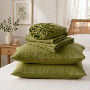 Puede incluir: Un juego de ropa de cama color verde oliva que incluye dos fundas de almohada rectangulares apiladas en la base, cubiertas por una sábana encimera doblada y una sábana bajera con bordes elásticos. La tela tiene un tejido texturizado visible.