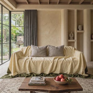 May include: A sofa covered with a light yellow throw blanket featuring a dark green border, two gray square throw pillows, and a wooden coffee table holding a stack of three books and a bowl filled with several red pomegranates.