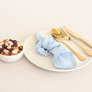 May include: A white plate with a light blue napkin, a gold fork, a gold knife, and a gold spoon. A white bowl filled with mixed nuts and red beans is on the left side of the image.
