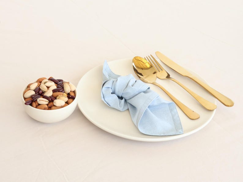 May include: A white plate with a light blue napkin, a gold fork, a gold knife, and a gold spoon. A white bowl filled with mixed nuts and red beans is on the left side of the image.