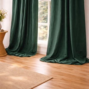 Puede incluir: Cortinas largas de color verde oscuro cuelgan frente a una ventana. Las cortinas están drapeadas y el suelo es de madera clara. Una alfombra tejida se encuentra frente a las cortinas. La ventana muestra el exterior.
