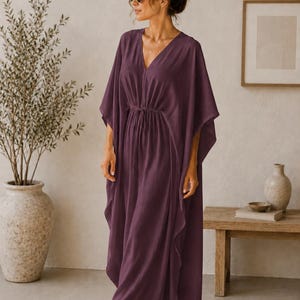 May include: A plum-colored, flowing caftan dress with a V-neckline and a drawstring waist. The dress has wide sleeves and a floor-length hem. The model is wearing sunglasses. The dress is made of a lightweight fabric, perfect for warm weather.