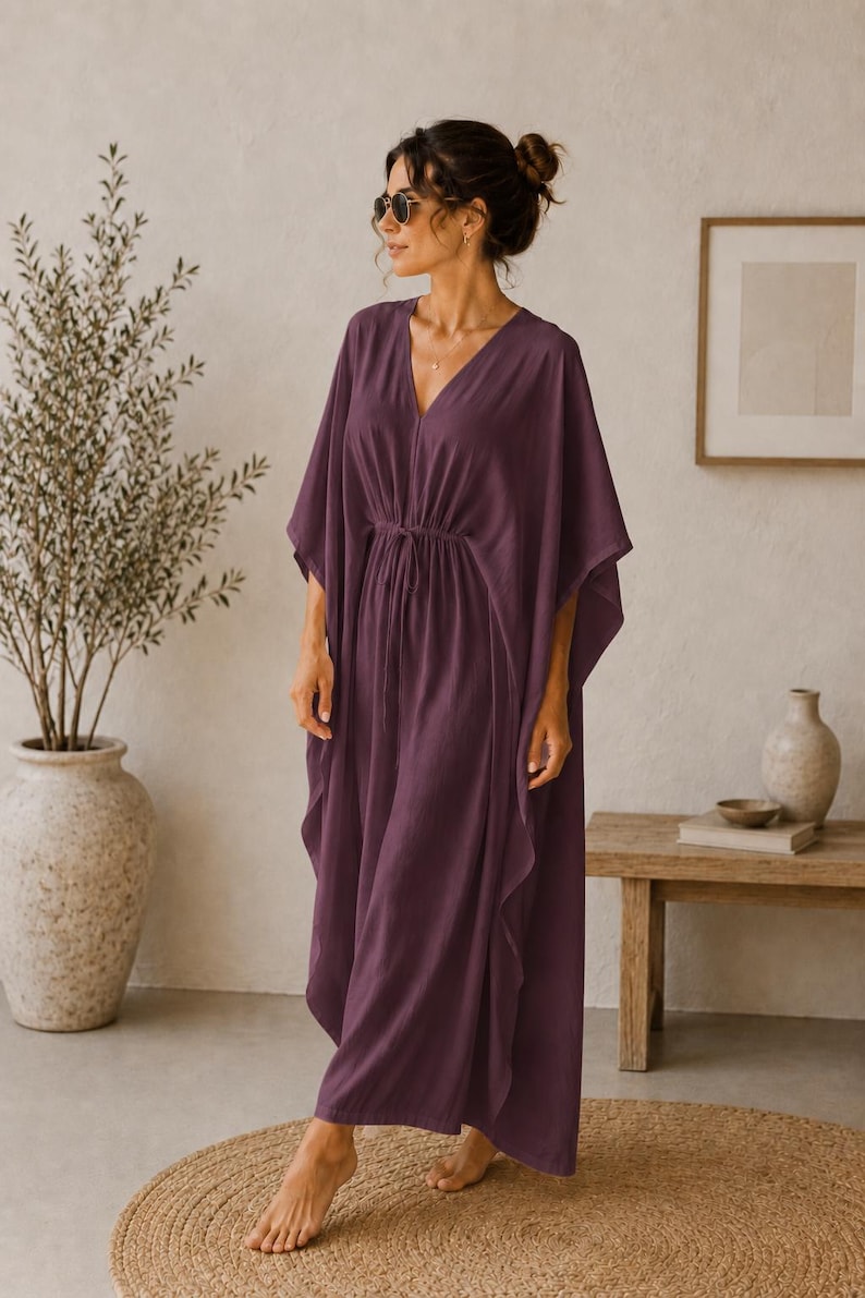 May include: A plum-colored, flowing caftan dress with a V-neckline and a drawstring waist. The dress has wide sleeves and a floor-length hem. The model is wearing sunglasses. The dress is made of a lightweight fabric, perfect for warm weather.