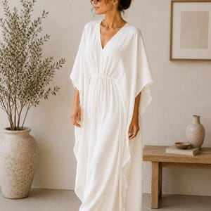May include: A white, flowing dress with a V-neck and wide sleeves. The dress has a cinched waist and falls to the floor. The woman is wearing sunglasses and a necklace. The background includes a plant in a large vase and a wooden bench.