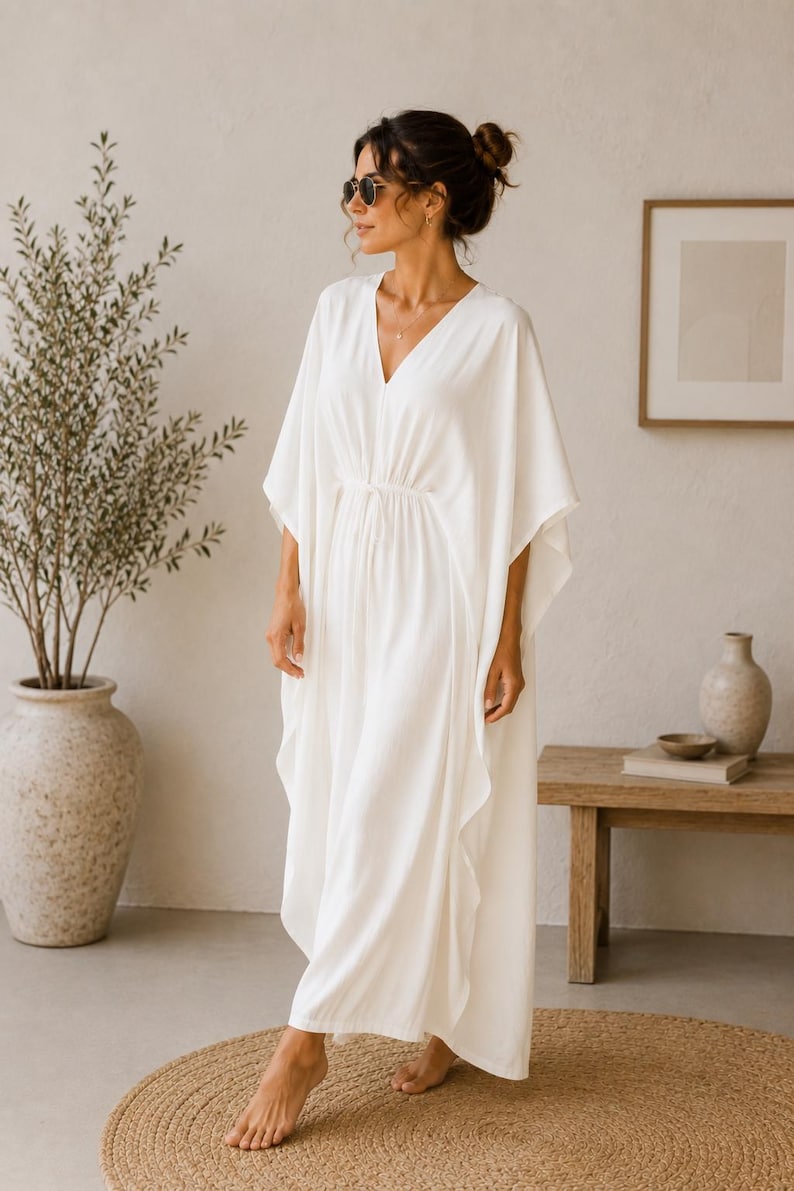 May include: A white, flowing dress with a V-neck and wide sleeves. The dress has a cinched waist and falls to the floor. The woman is wearing sunglasses and a necklace. The background includes a plant in a large vase and a wooden bench.