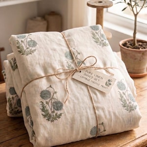 Può includere: Un set copripiumino in lino bianco sporco piegato con una stampa di cardi verdi e blu, legato con spago marrone e un'etichetta di carta che riporta la scritta PURE LINEN DUVET SET - COTTAGE THISTLE PRINT. Il tessuto ha una trama visibile.
