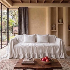 May include: A white sofa covered with a long, ruffled fabric slipcover, featuring two matching white square throw pillows. In front of the sofa sits a rectangular wooden coffee table holding a stack of books and a bowl filled with red pomegranates.