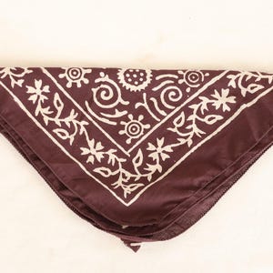 May include: A maroon bandana with a white floral and geometric pattern. The bandana is folded in a triangle shape, with the design framing the edges. The fabric appears to be soft and smooth.