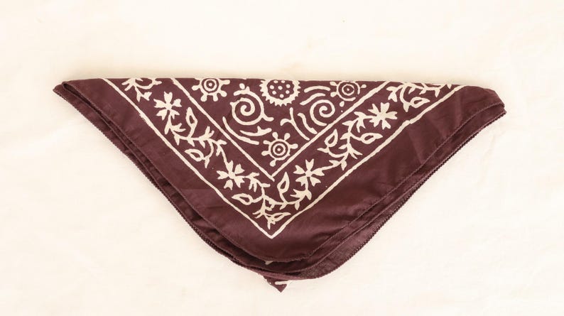 May include: A maroon bandana with a white floral and geometric pattern. The bandana is folded in a triangle shape, with the design framing the edges. The fabric appears to be soft and smooth.