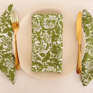 May include: Three white napkins with a green floral pattern are folded and placed on a white tablecloth. A gold fork and knife are placed on a white plate.