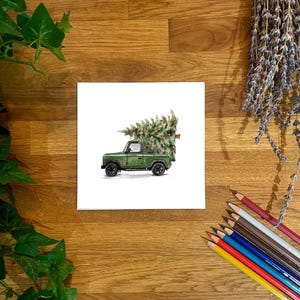May include: A watercolour illustration depicts a green pickup truck transporting a Christmas tree. The truck has black wheels and a white cab. The tree is a vibrant green with red decorations. The artwork is on a white square, placed on a wooden surface.