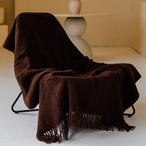 Op de afbeelding: Een donkerbruine, gefranjerde sprei over een zwarte metalen stoel. De sprei heeft een textuur. De achtergrond toont een neutraal getinte muur met een decoratieve nis en een kleine tafel. De kamer heeft een minimalistisch ontwerp.