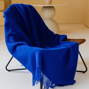 Pode incluir: Um cobertor azul vibrante com franjas, drapeado sobre uma cadeira moderna. A cadeira tem uma estrutura de metal preta e um assento tecido. A cor rica do cobertor contrasta com os tons neutros da sala, criando um ambiente acolhedor.