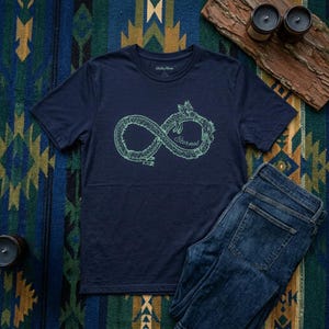 Puede incluir: Camiseta azul marino con un símbolo de infinito verde claro que representa un dragón y la palabra "Eternal". La camiseta se muestra con unos vaqueros azules. El diseño está impreso en la parte delantera de la camiseta.