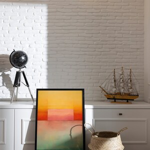 Peut inclure: Une peinture abstraite encadr&eacute;e aux couleurs jaune, orange, rose et verte est accroch&eacute;e &agrave; un mur en briques blanches au-dessus d'une armoire blanche. Un globe terrestre sur un tr&eacute;pied se trouve sur l'armoire &agrave; gauche de la peinture. Un petit voilier en bois se trouve sur l'armoire &agrave; droite de la peinture. Un panier en osier se trouve sur le sol devant l'armoire.