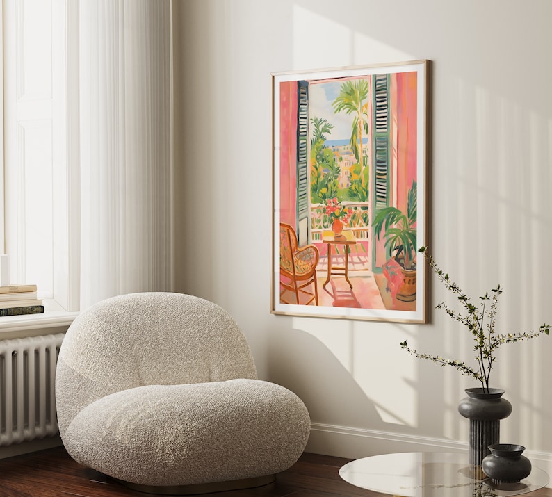 May include: A framed print of a painting depicting a pink room with a window overlooking a cityscape. The window has green shutters and a balcony with a small table and chairs. There are plants in pots on the balcony and a vase of flowers on the table.