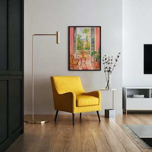 May include: A yellow armchair sits in a living room with a painting of a balcony overlooking a city. The painting is framed in black and hangs on a white wall. A floor lamp with a gold base and a gold arm is in the background. A small table with a vase of flowers sits next to the armchair.