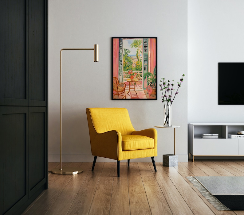 May include: A yellow armchair sits in a living room with a painting of a balcony overlooking a city. The painting is framed in black and hangs on a white wall. A floor lamp with a gold base and a gold arm is in the background. A small table with a vase of flowers sits next to the armchair.