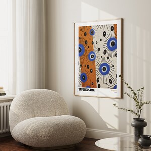 May include: A framed print of a colorful abstract artwork by Yayoi Kusama. The artwork features a pattern of blue and orange eyes with black outlines on a white background. The print is framed in a light wood frame and is hanging on a white wall.