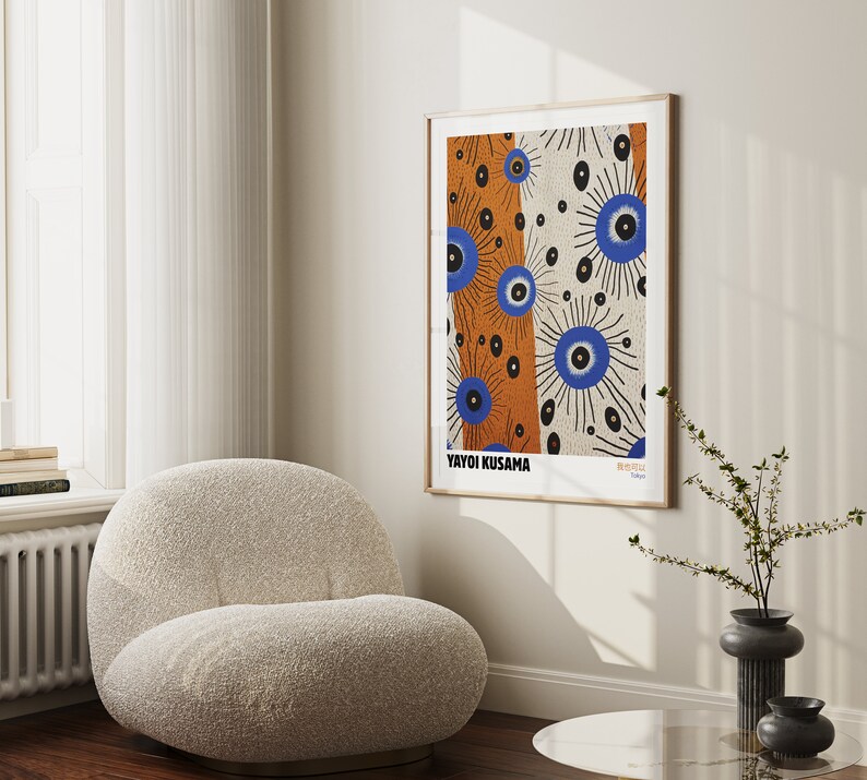 May include: A framed print of a colorful abstract artwork by Yayoi Kusama. The artwork features a pattern of blue and orange eyes with black outlines on a white background. The print is framed in a light wood frame and is hanging on a white wall.