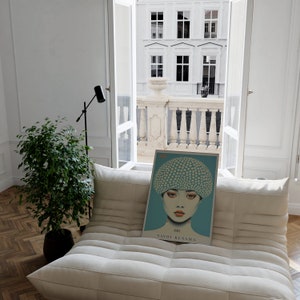 May include: A framed art print displaying a portrait of a woman with a polka-dotted hat on a teal background. The print rests on a white sofa in a bright room, with a view of a European-style building visible through the window.