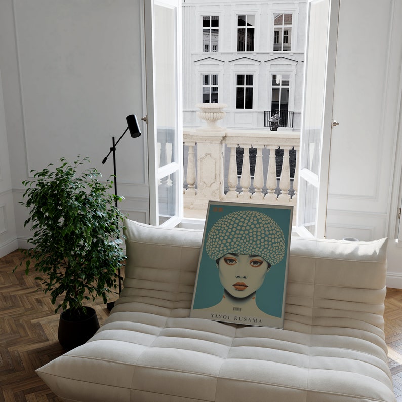 May include: A framed art print displaying a portrait of a woman with a polka-dotted hat on a teal background. The print rests on a white sofa in a bright room, with a view of a European-style building visible through the window.