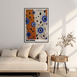 May include: A white couch with three pillows and a wooden side table with a vase of dried branches. A framed poster with a colorful abstract design by Yayoi Kusama hangs on the wall above the couch. The poster features a repeating pattern of blue and orange circles with black lines.
