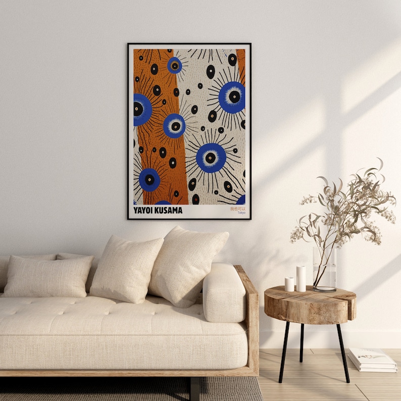 May include: A white couch with three pillows and a wooden side table with a vase of dried branches. A framed poster with a colorful abstract design by Yayoi Kusama hangs on the wall above the couch. The poster features a repeating pattern of blue and orange circles with black lines.