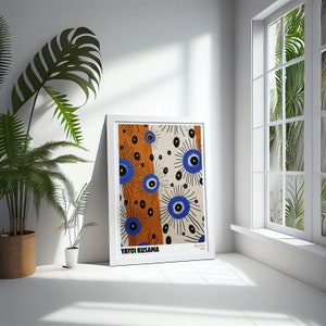 May include: A white framed print of an abstract art piece by Yayoi Kusama. The print features a repeating pattern of blue and black eyes on a white and orange background. The artist's name is printed at the bottom of the print.