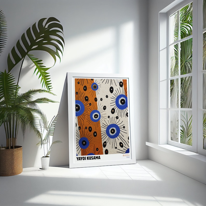 May include: A white framed print of an abstract art piece by Yayoi Kusama. The print features a repeating pattern of blue and black eyes on a white and orange background. The artist's name is printed at the bottom of the print.