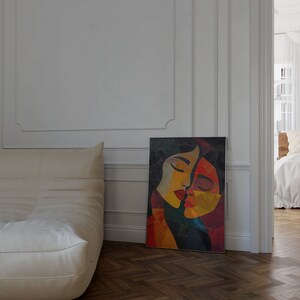May include: Abstract painting of two faces in a close embrace, with a colourful palette of red, yellow, orange, and blue. The painting is framed in a black frame and is hanging on a white wall.