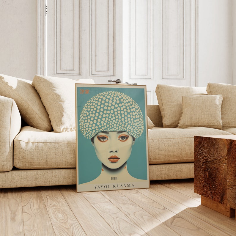 May include: A framed art print of a woman with a dotted head covering and red lips, by Yayoi Kusama. The artwork has a teal background and is displayed on a wooden floor next to a beige sofa. The print has the artist's name.