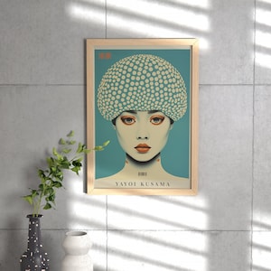 May include: A framed art print of a woman with a dotted head covering and orange makeup. The artwork, set against a teal background, is signed "YAYOI KUSAMA" and is in a light wood frame. The image is displayed on a concrete wall.