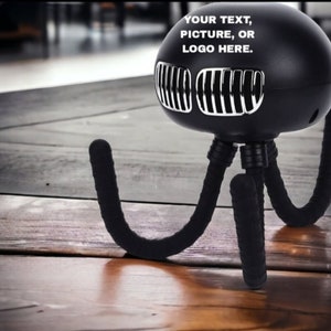 May include: Black octopus-shaped phone holder with three flexible legs. The holder has a silver grill on the front and the text "YOUR TEXT, PICTURE, OR LOGO HERE." is printed on the top.