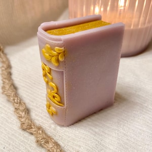 May include: A lavender-colored book-shaped soap with gold-colored gilded edges and decorative accents. The soap has a glittery finish and is set against a soft, neutral background. A decorative item for the bathroom.