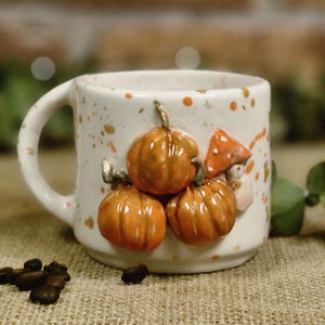 Peut inclure: Une tasse en céramique blanche avec une anse, ornée de trois citrouilles oranges et d'un champignon. La tasse présente des mouchetures marron et est posée devant un mur de briques flou. Des grains de café sont éparpillés au premier plan.