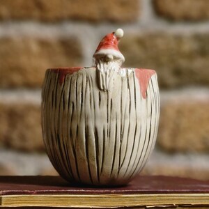 May include: A ceramic Santa Claus figurine peeks over the edge of a textured, cream-colored bowl with vertical lines. The bowl sits atop two antique books with worn, maroon covers. The background features a brick wall.