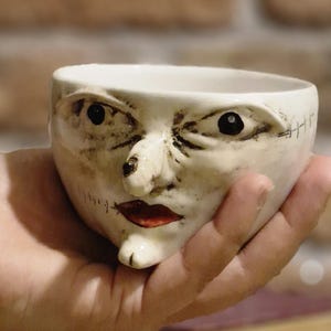 Può includere: Una tazza in ceramica bianca con un design a volto, labbra rosse e dettagli cuciti. La tazza è tenuta in mano, con una pila di libri e chicchi di caffè sullo sfondo. Un braccialetto verde e argento è sul polso.
