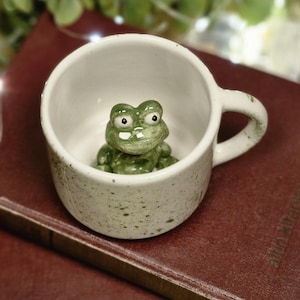 May include: A small, speckled white ceramic mug with a green frog figurine inside. The mug is placed on top of two maroon books. The background features greenery and fairy lights, creating a cosy, whimsical scene.