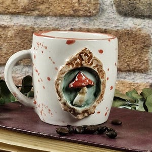 May include: White ceramic mug with a red and brown mushroom design. The mug has a handle and is decorated with red paint splatters. The mushroom is set in a decorative frame. Coffee beans are scattered near the mug.