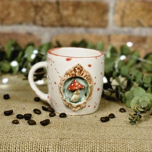 May include: A white ceramic mug with a red rim and red speckles. The mug features a 3D mushroom design in a decorative frame. Coffee beans are scattered around the mug on a burlap surface.
