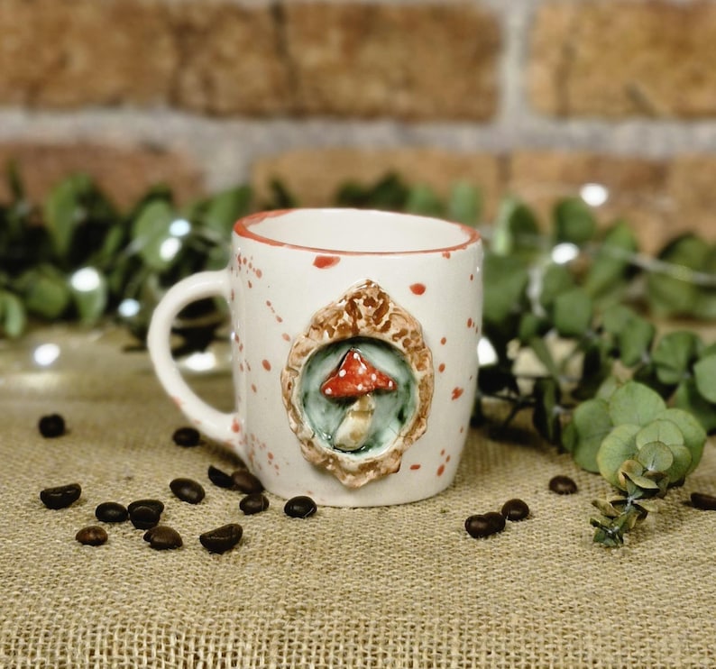 May include: A white ceramic mug with a red rim and red speckles. The mug features a 3D mushroom design in a decorative frame. Coffee beans are scattered around the mug on a burlap surface.