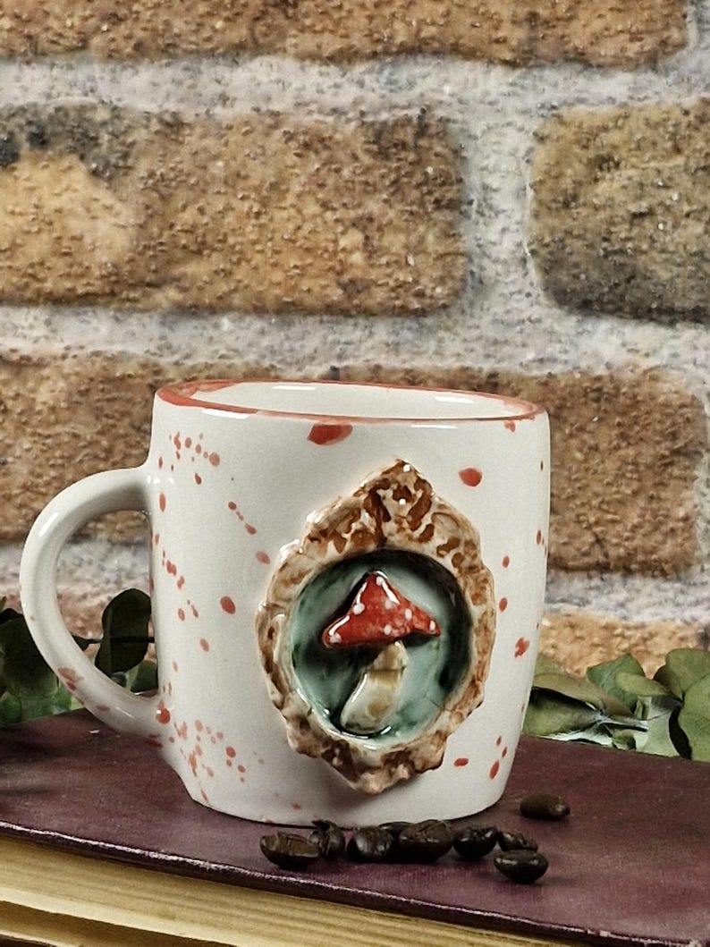 May include: White ceramic mug with a red and brown mushroom design. The mug has a handle and is decorated with red paint splatters. The mushroom is set in a decorative frame. Coffee beans are scattered near the mug.