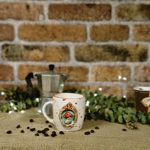 May include: A white ceramic mug with a red rim and a mushroom design. The mug has a handle and is decorated with a framed mushroom illustration. Coffee beans are scattered around the mug, with a brick wall in the background.